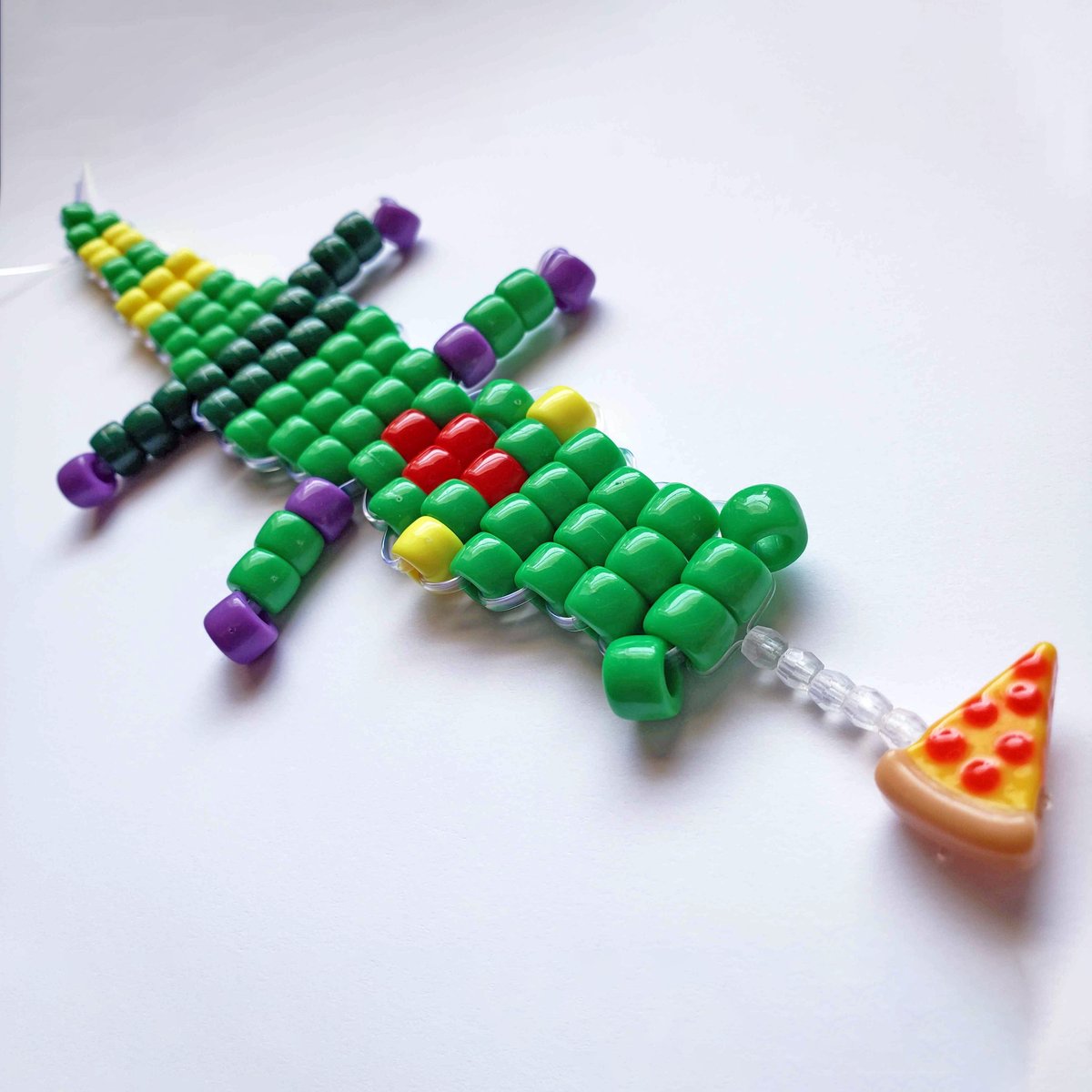 a beadwork alligator figure, with red hair, dark green pants, and a yellow-striped tail. He has a slice of pizza hanging down from his mouth.