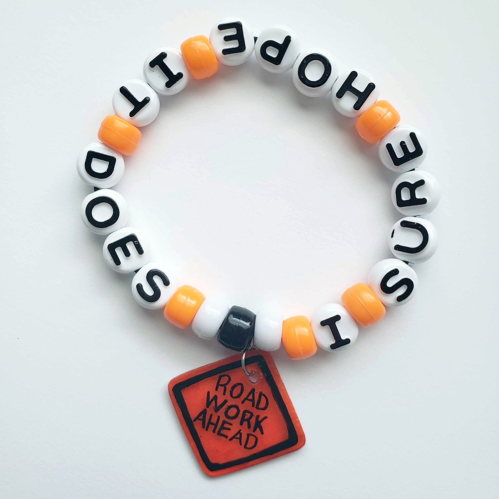 a black, white and bright orange kandi-style bracelet. It has a charm that looks like an orange sign reading 'Road Work Ahead,' while the beads on the bracelet continue with 'I sure hope it does.'