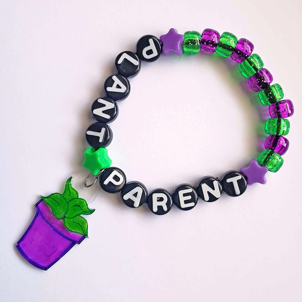 a purple and green, glittery beaded bracelet that says 'Plant Parent.' A plastic charm of a green plant in a purple pot hangs from the middle of the words.