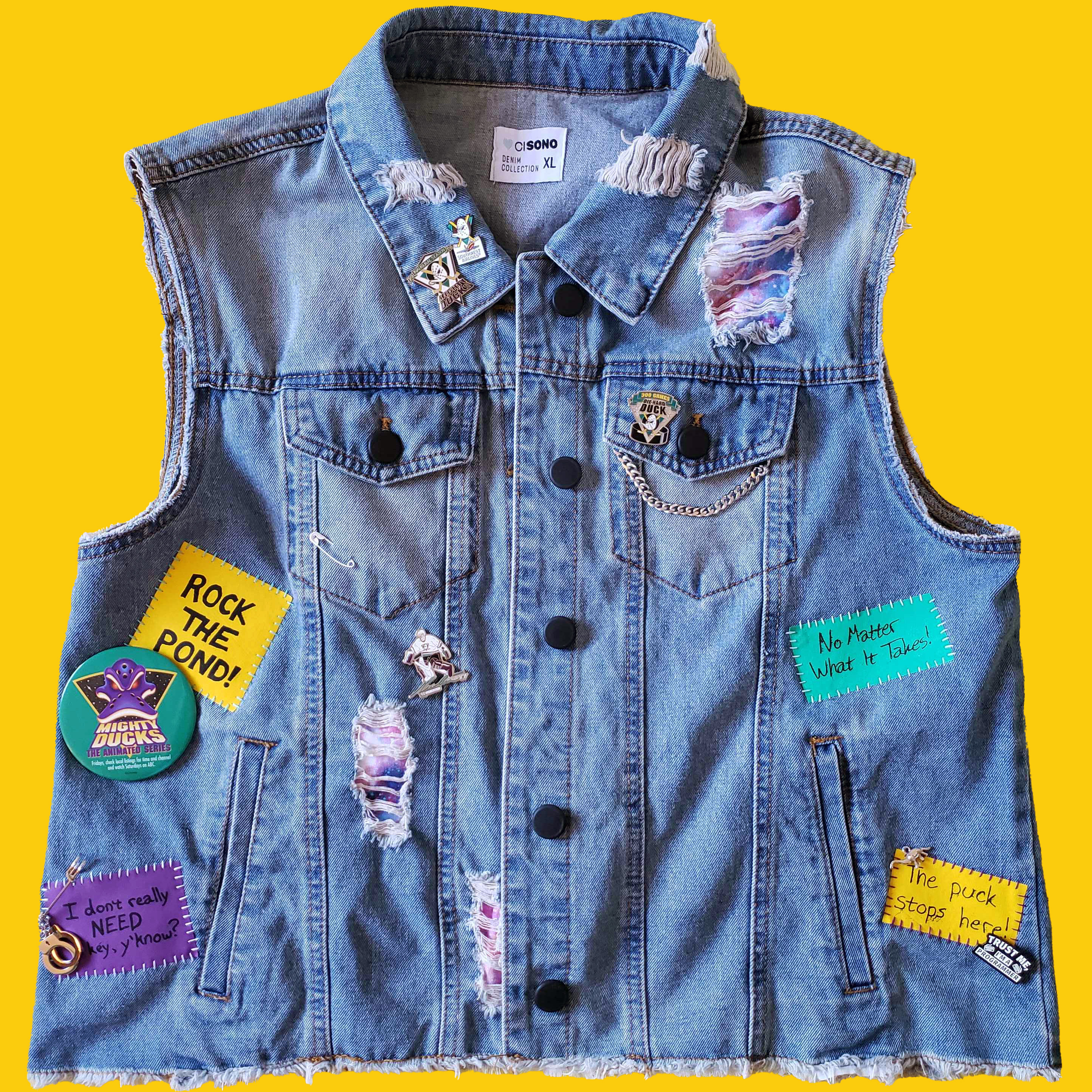 A distressed denim vest with black buttons, covered in pins, patches and charms for both Mighty Ducks the real hockey team and Mighty Ducks the cartoon. There are multiple rips in the vest, behind which a galaxy print fabric can be seen.