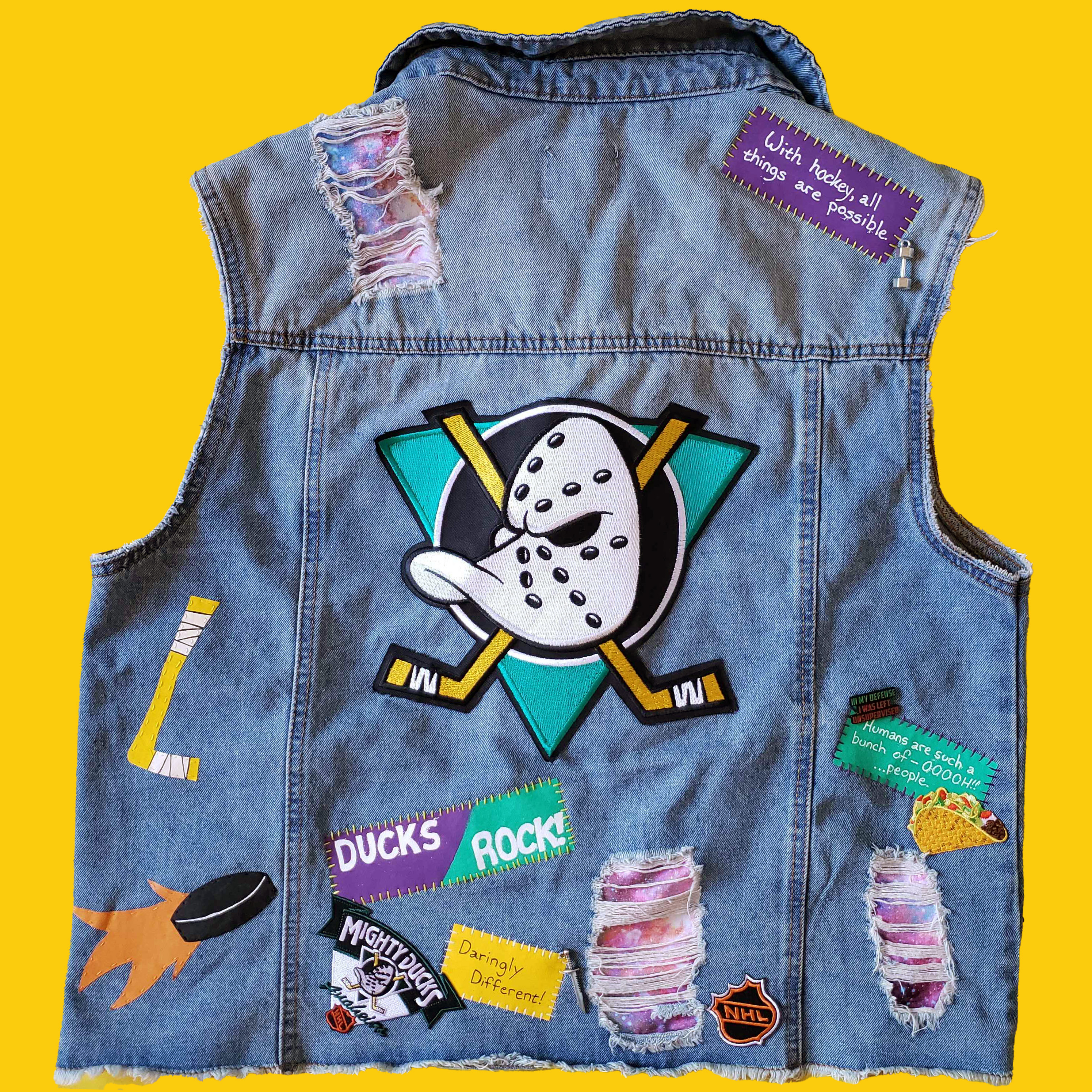 The back of a distressed denim vest, covered in pins, patches and charms for both Mighty Ducks the real hockey team and Mighty Ducks the cartoon. There are multiple rips in the vest, behind which a galaxy print fabric can be seen.