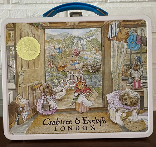 a white tin lunchbox with a blue plastic handle, with a large Beatrix Potter illustration featuring many of her characters, most notably Peter Rabbit