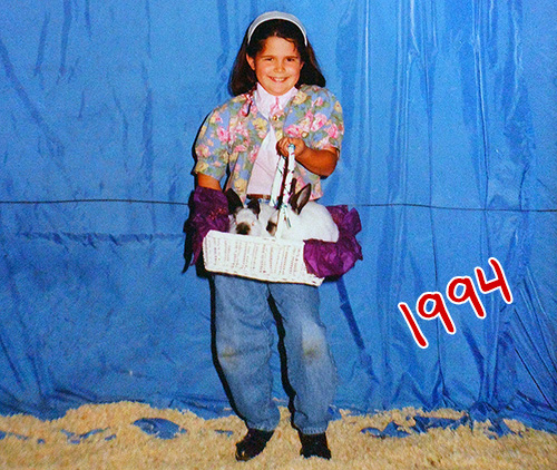 a smiling young girl in blue jeans, a pastel floral top, and long brown hair held back by a headband, holding a white basket lined with sparkly purple tissue paper and itself holding two mid-size white rabbits with dark snouts and ears
